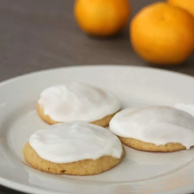 3 frosted orange cookies on a white plate with fresh oranges in the background.