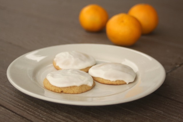 3 frosted orange cookies on a white plate with 2 fresh oranges in the background.