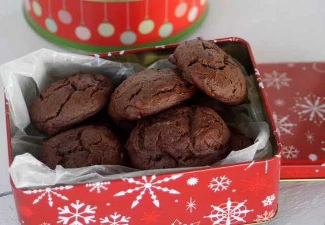 Chocolate cheesecake cookies in a red tin decorated with white snowflakes.