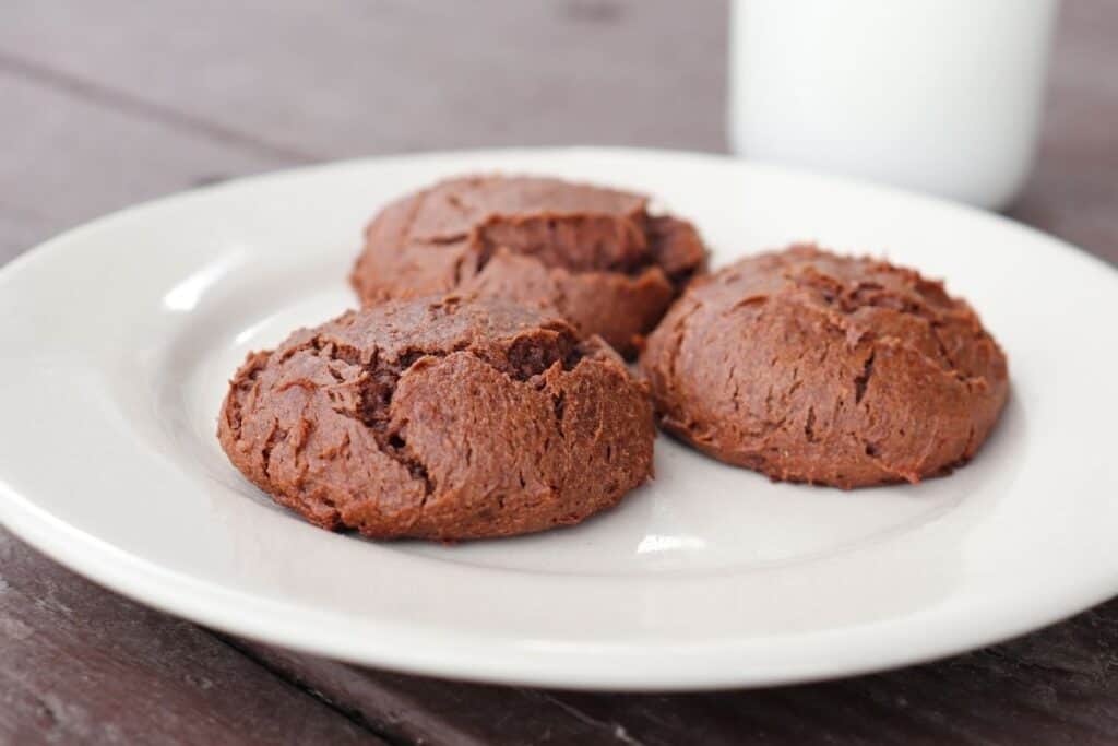 3 chocolate cheesecakes cookies on a white plate with a glass of milk in the background.