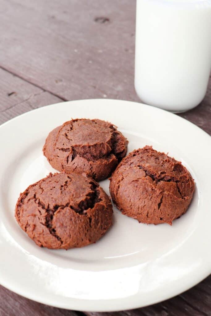 3 chocolate cheesecakes cookies on a white plate as seen from above with a glass of milk in the background.