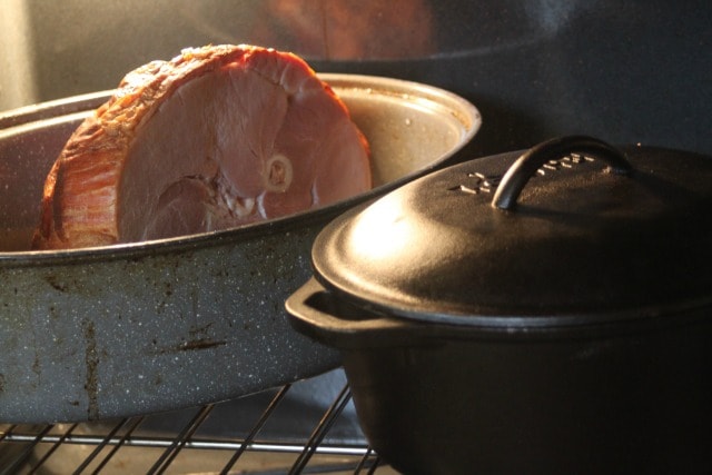 A ham in roasting sitting behind covered cast iron dutch oven in an oven.
