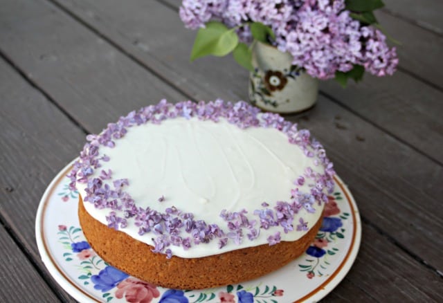 A lilac honey cake on a cake plate with a vase of fresh lilac branches in the background.