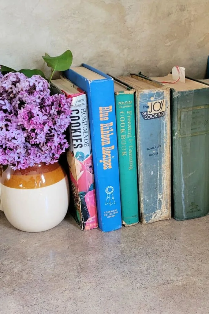 A row of vintage cookbooks sits on a shaelf with a vase full of lilac flowers.