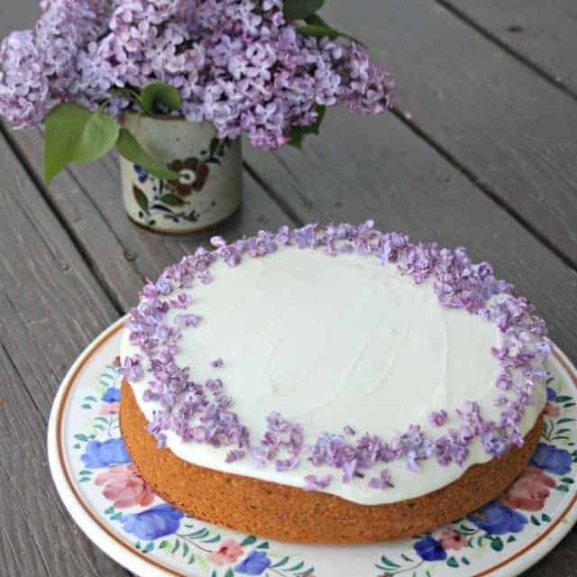 A frosted cake decorated with lilac flowers on a floral cake plate with a vase of fresh lilacs in the background.