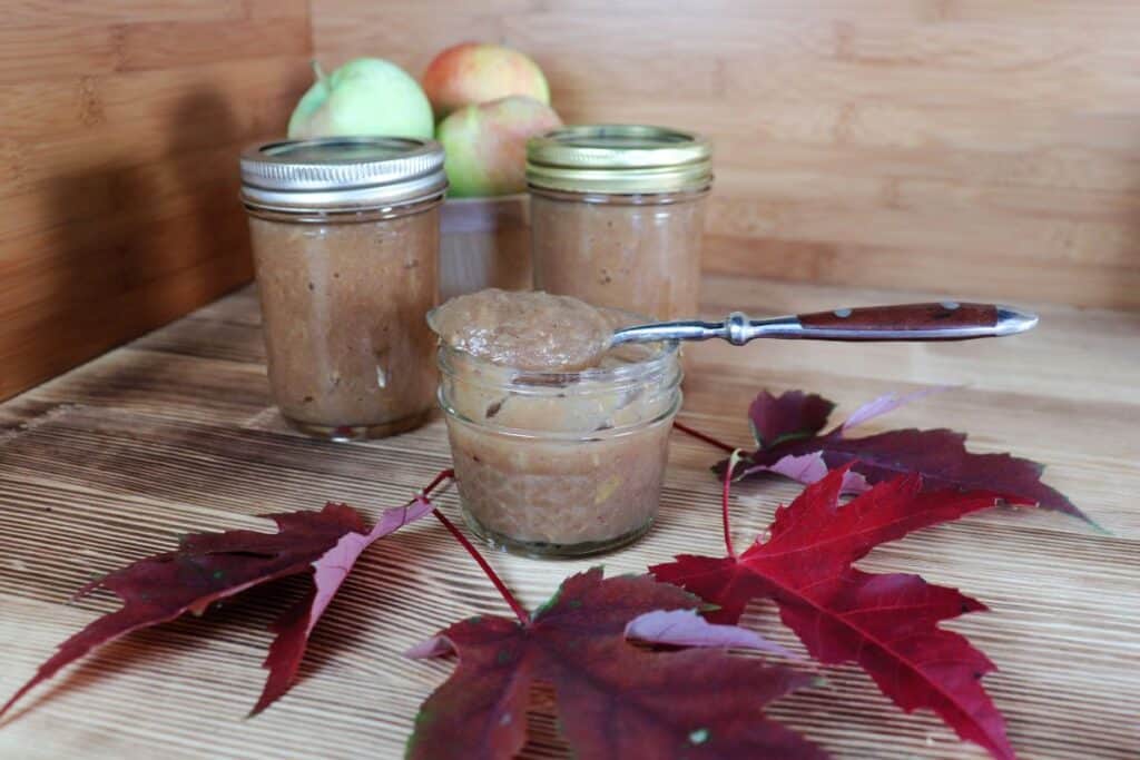 A spoon with apple butter sits on top of an open jar of apple butter. In front of the jars are some red maple leaves. Behind the jar are more sealed jars of apple butter and a basket of fresh apples.