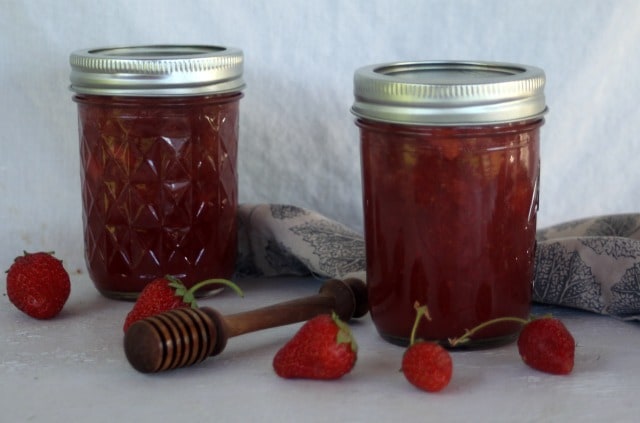 Two jars of strawberry jam on a table with fresh strawberries, a honey dipper, and cloth napkin