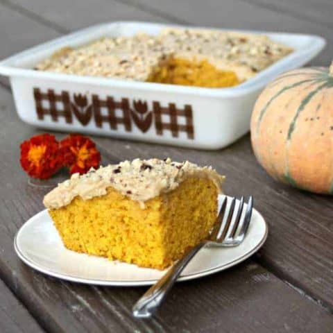 A slice of frosted cake on a white plate with a fork, the pan of remaining cake in the background with a pumpkin.