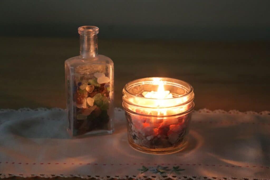 A lit candle in a jar sits on a table runner with an antique bottle full of peblles next to it.
