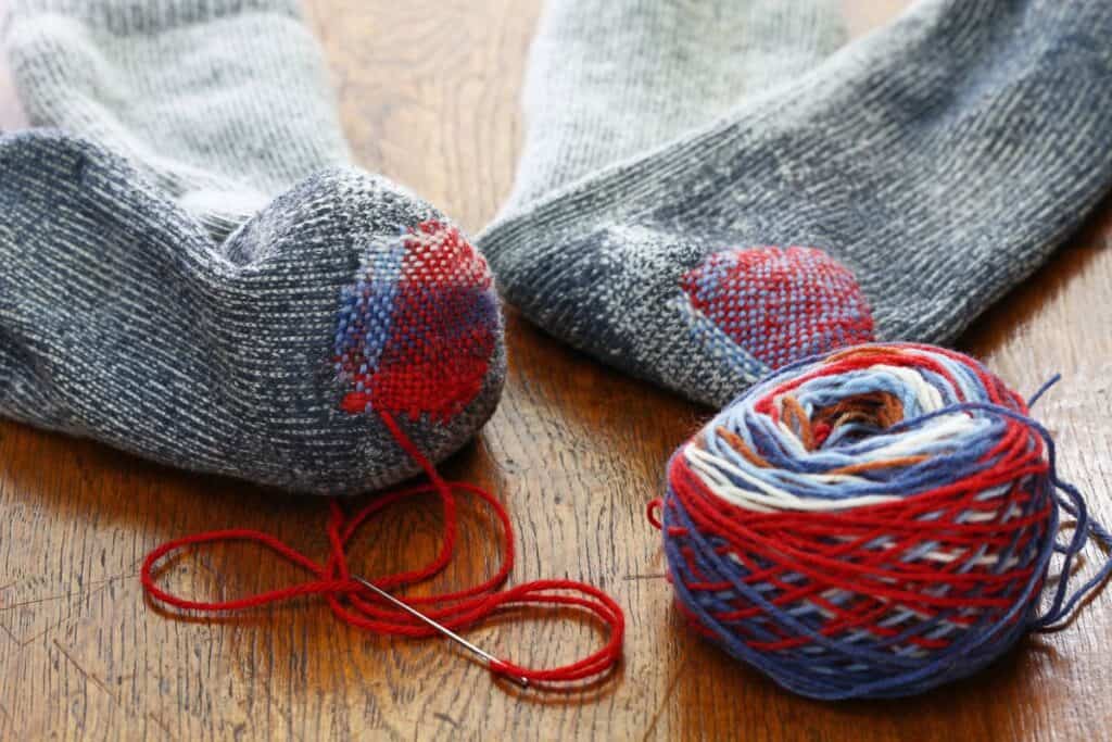2 socks sit on a table with a ball of yarn and a needle. The socks have had holes darned.