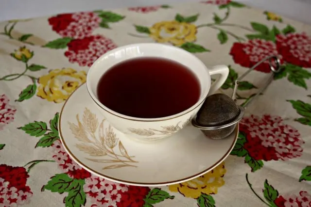 A cup of hibiscus tea in a cup sitting on a saucer with a tea ball on the plate. 