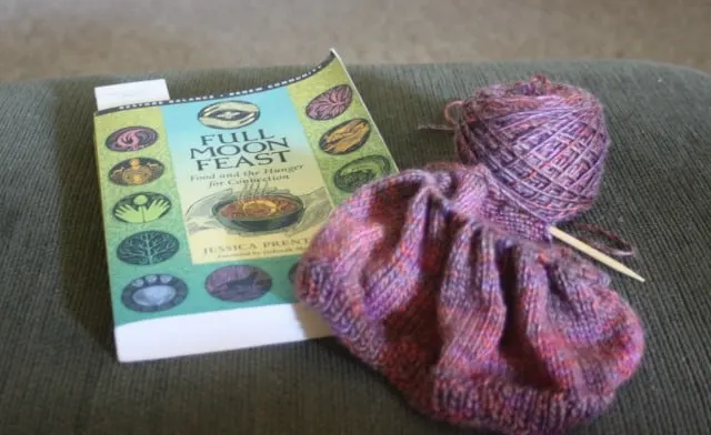 a book and knitting project with needles and ball of yarn on ottoman.