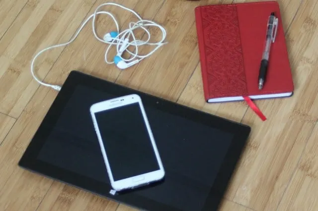 Smartphone stacked on top of a tablet connected to ear buds sitting near a journal with a pen.