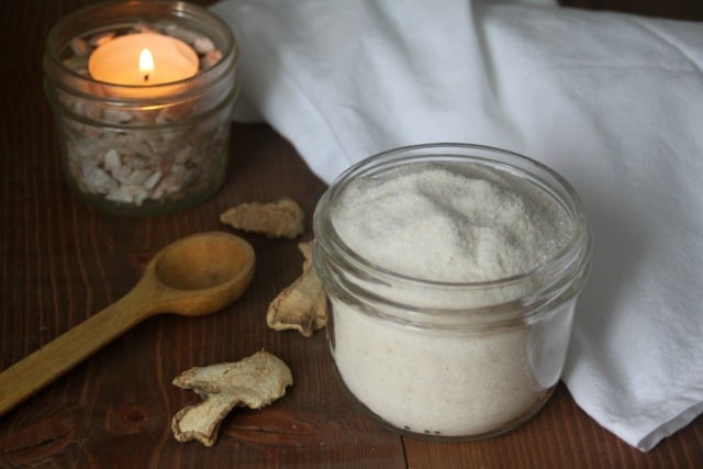 Ginger detox bath salts in a jar surrounded by a candle, wooden spoon, towel, and dried herbs on a table. 