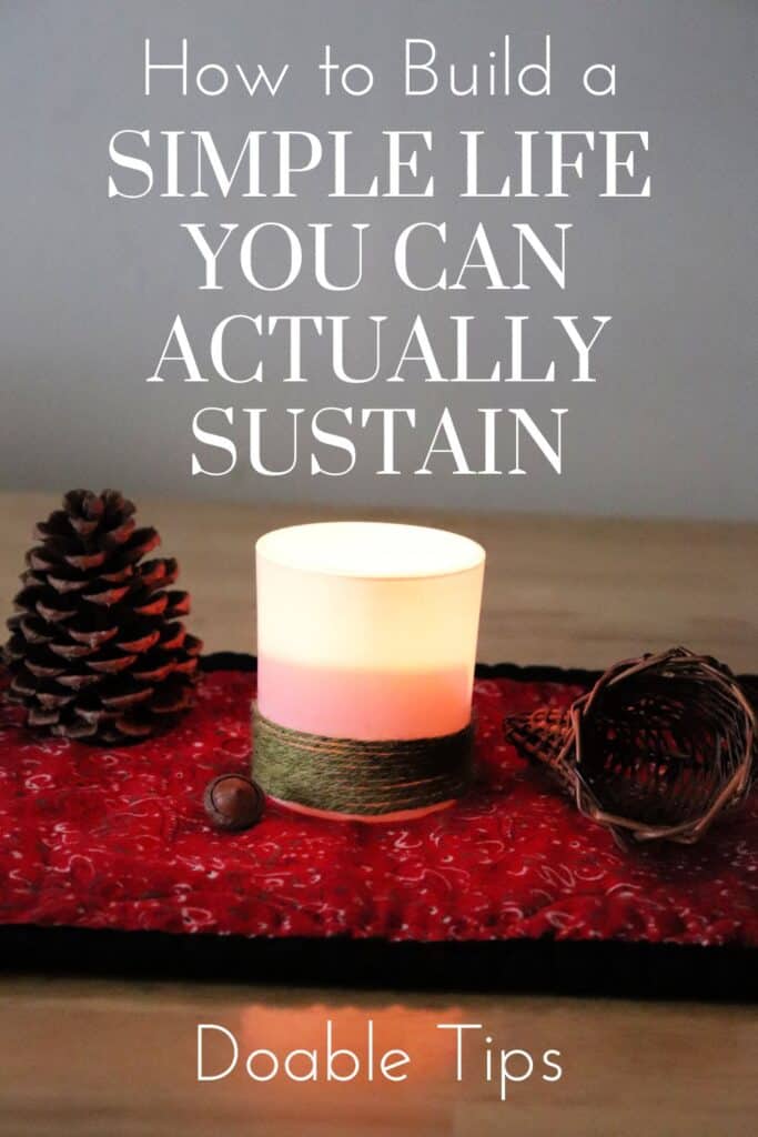 An illuminated candle holder sits on a table runner with a small acorn, a pinecone, and a small basket. Text overlay reads: How to  Build a Simple Life You Can Actually Sustain. 
