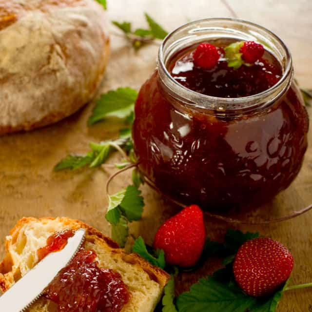 Strawberry jam in an open jar, toast spread with jam and fresh strawberries on a table.