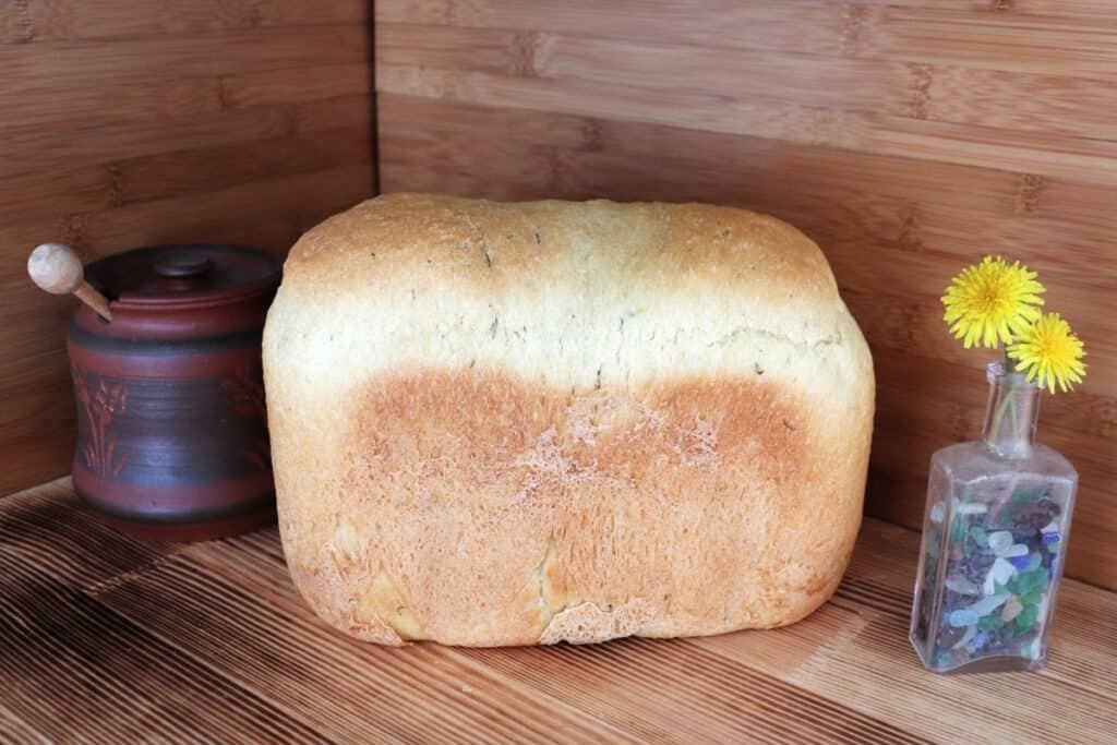 A loaf of bread sits on the counter between a small vase holding dandelion flowers and a clay honey pot. 