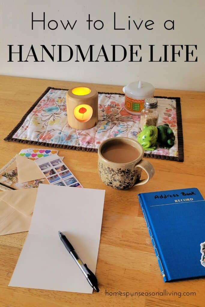 An open notecard with a pen sits on a table next to an address book. A cup of coffee, candle, and jar sit on placemat in the center of the table. Text overlay reads: how to live a handmade life.