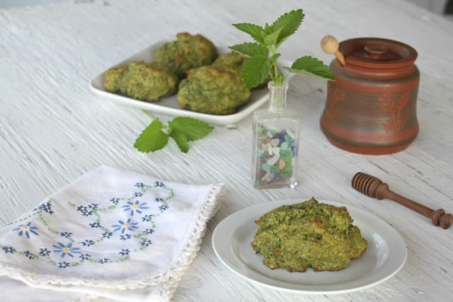 A lemon balm drop biscuit on a plate sitting next to a napkin with a bottle full of fresh lemon balm leaves, a honey jar, a honey dipper, and a platter of more biscuits in the background.