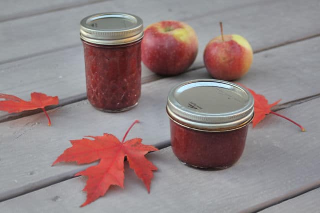 Jars of cranberry apple butter on a table with red maple leaves and fresh apples.