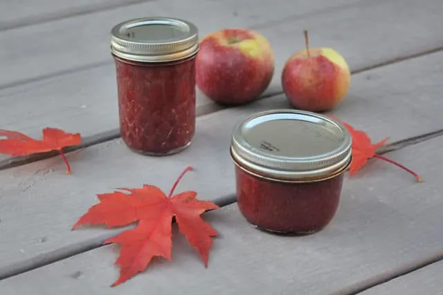 Jars of cranberry apple butter on a table with red maple leaves and fresh apples. 