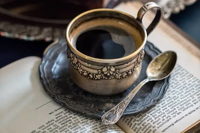 A cup of coffee with teaspoon sitting on a saucer on open book.