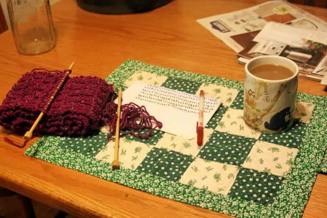 A scarf on knitting needles next to a cup of coffee on table. 