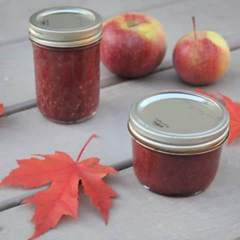 Jars of cranberry apple butter on a table with maple leaves and fresh apples.