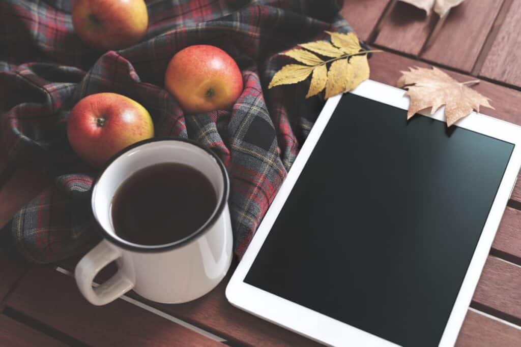 An e-tablet sits on a table next to a cup of coffee. A plaid cloth sits on the table with fresh apples on top of it. 