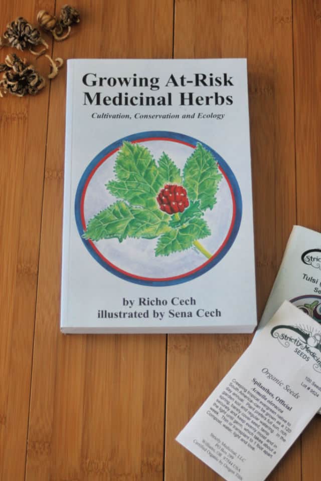 The book Growing At-Risk Medicinal Herbs sitting on a table with seed packets and loose seeds.