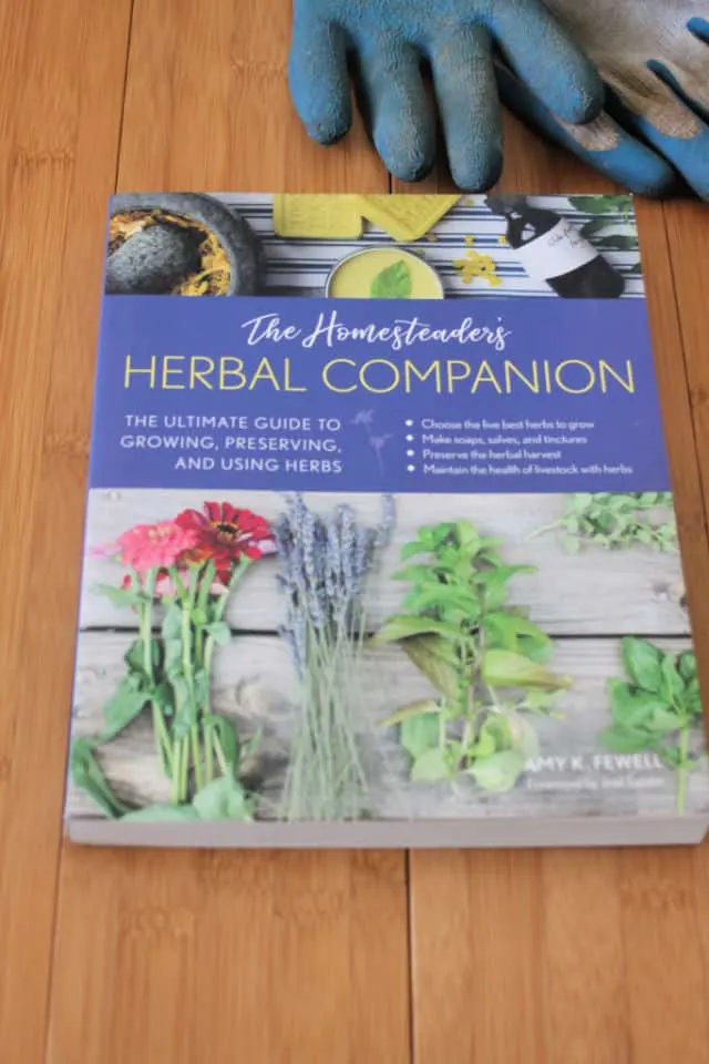 The book The Homesteader's Herbal Companion on a table with a pair of gardening gloves.