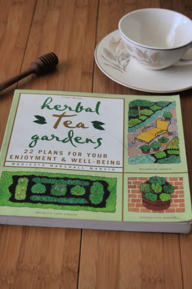 The book Herbal Tea Gardens on a table with honey dipper and tea cup on a saucer.