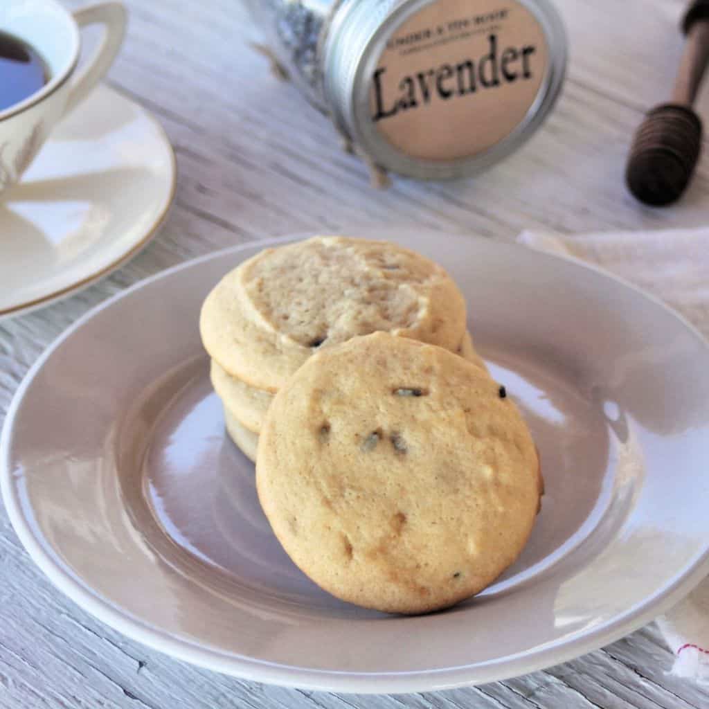 Lavender cookies on a white plate with a tea cup and jar of lavender buds in the background.