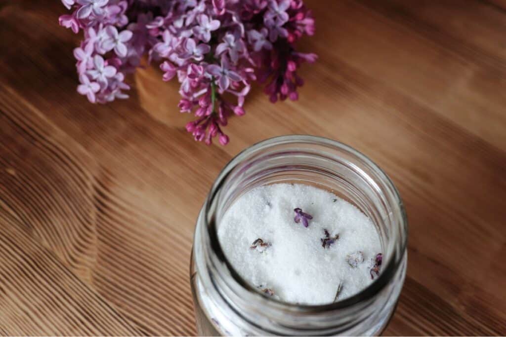 An open jar as seen from above contains sugar and dried lilac blossoms. Fresh branches of lilacs sits behind the jar. 