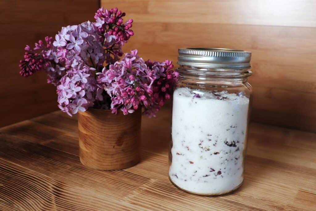 An open canning jar full of sugar and dried lilac blossoms sit on a table in front of a vase of fresh lilac branches.