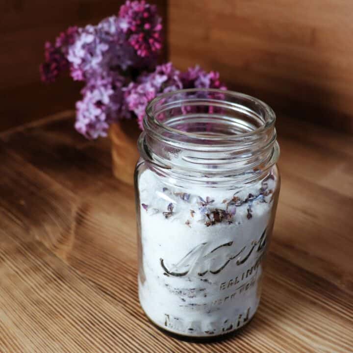 An open canning jar full of sugar and dried lilac blossoms sit on a table in front of a vase of fresh lilac branches.