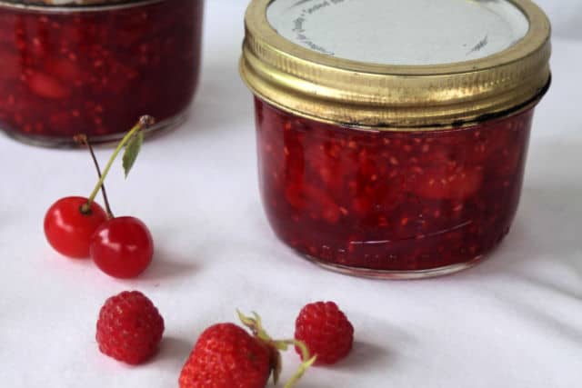 Jars of brandied cherry berry preserves on a white table surrounded by fresh berries and cherries.