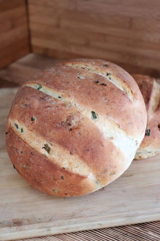 2 loaves of bread showing bits of green chives and potato skins on the crust sit on a board.