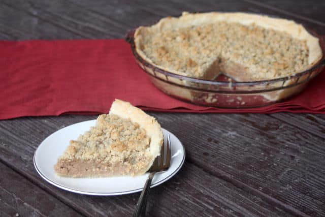 A slice of apple butter pie on a plate with a fork sitting in front of a whole pie on a red table runner.