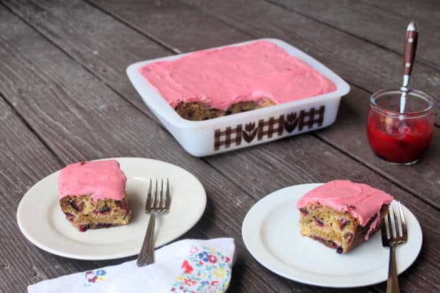 Two slices of plum cake with plum jam frosting on plates with forks sitting in front of whole cake and jar of plum jam.