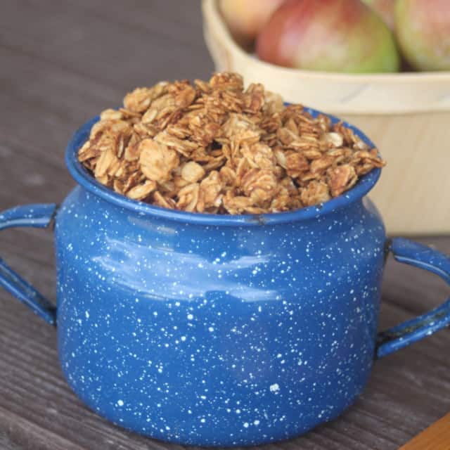 Applesauce Peanut Butter Granola piled into a blue tin cup with a basket of fresh apples behind it.