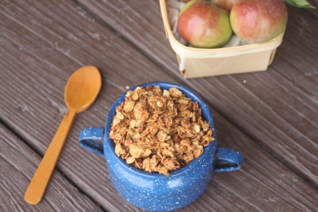 Applesauce peanut butter granola piled into a blue tin cup, a wooden spoon sitting to the left, a basket of fresh apples behind it.