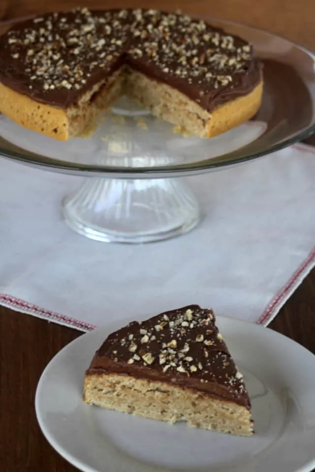 A slice of honey cake with chocolate honey frosting on a plate sitting in front of the entire cake on a cake pedestal. 