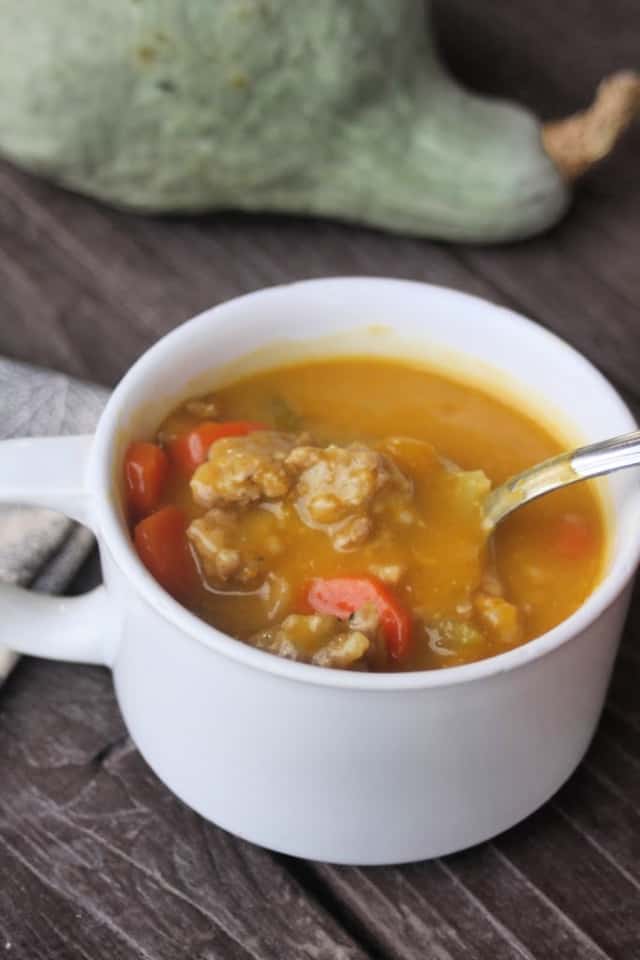 Hubbard squash sausage soup in a white cup with a spoon sitting in front of a hubbard squash. 