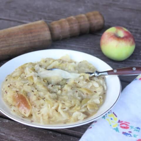 Cooked apple sauerkraut side dish in a bowl with a spoon sitting next to a napkin with a sauerkraut pounder and apple behind it.