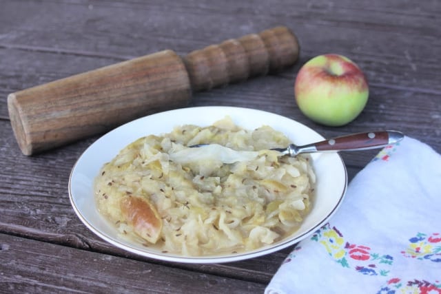 Cooked apple sauerkraut side dish in a bowl with a spoon sitting next to a napkin with a sauerkraut pounder and apple behind it.