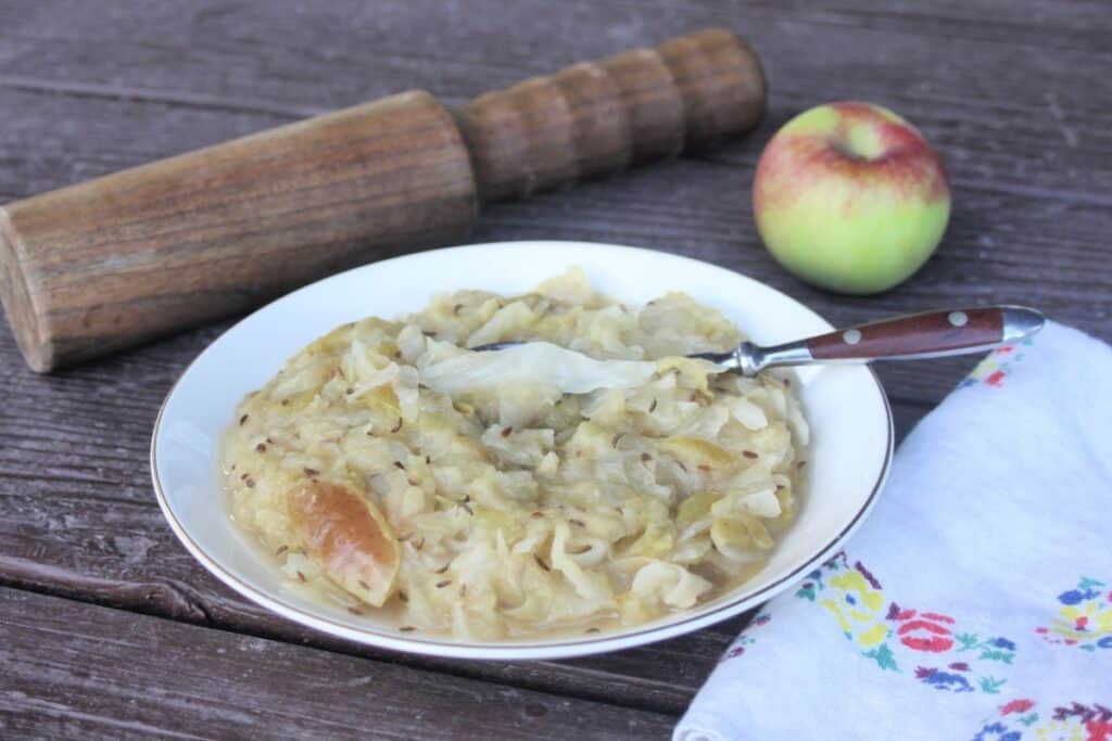 A bowl of cooked sauerkraut and apples with a spoon in it. A wooden mallet, fresh apple, and napkin surround the bowl.