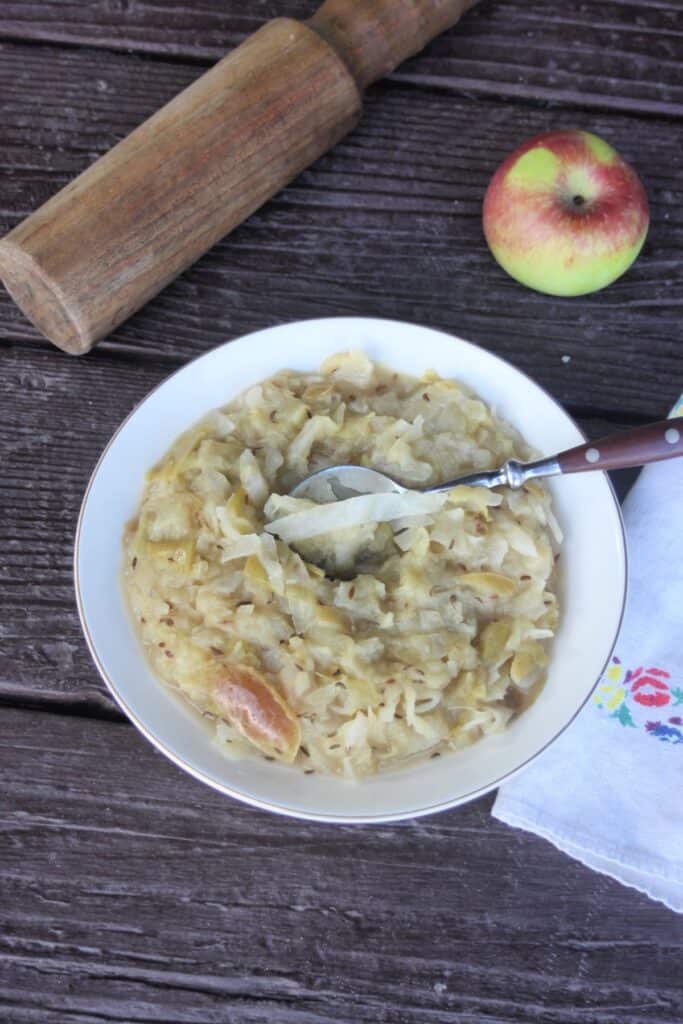 A bowl of cooked sauerkraut and apples with a spoon in it as seen from above. A wooden mallet, fresh apple, and napkin surround the bowl.