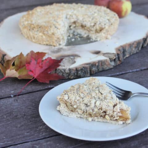 A slice of gluten-free apple tart on a plate with a fork sitting front of the entire tart sitting on a slice of wood.