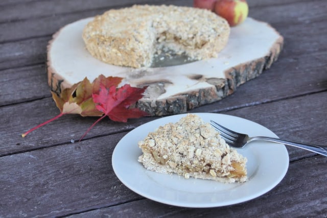 A slice of gluten free apple tart on a plate with entire tart on a wood slice behind it.